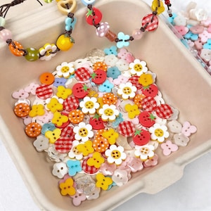 May include: A collection of colorful craft buttons in various shapes and sizes, including flowers, strawberries, and gingham patterns. The buttons are in a light brown container, with a few beaded bracelets in the background.