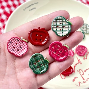 May include: A collection of apple-shaped buttons in various colors and patterns. The buttons include red, green, and pink with gingham, plaid, and solid designs. Each button has a small green stem and buttonholes for sewing.