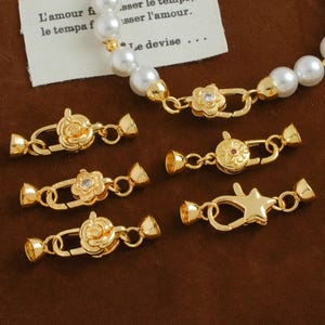 Può includere: Chiusure per gioielli dorate in vari disegni floreali e a stella. Le chiusure sono mostrate con una collana di perle e una piccola etichetta con testo in francese. Le chiusure sono progettate per collegare i gioielli.