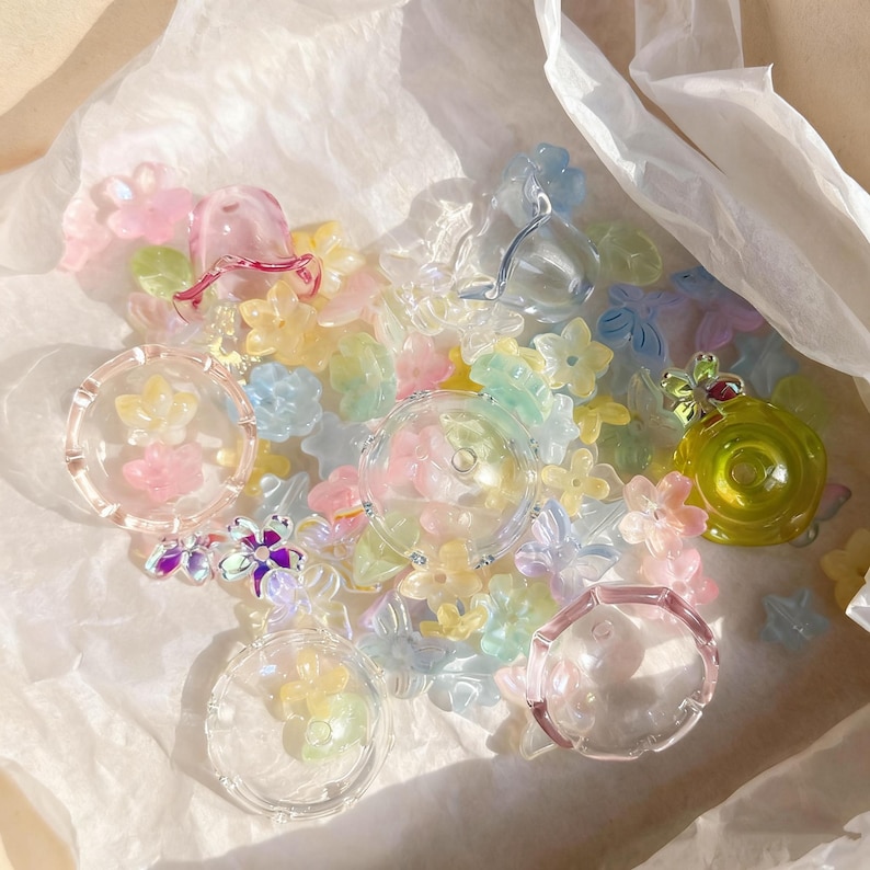 May include: A selection of pastel-coloured, translucent floral and butterfly-shaped beads and charms. Several clear, round, decorative pieces with flower accents are also present. The items are scattered on a white, crinkled surface.