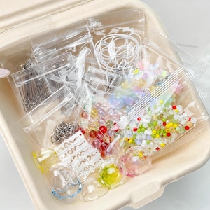May include: A craft kit with assorted beads, charms, and findings in clear plastic bags. The kit includes colourful beads, small metal pins, and clear glass-like components. The items are arranged in a light brown container.