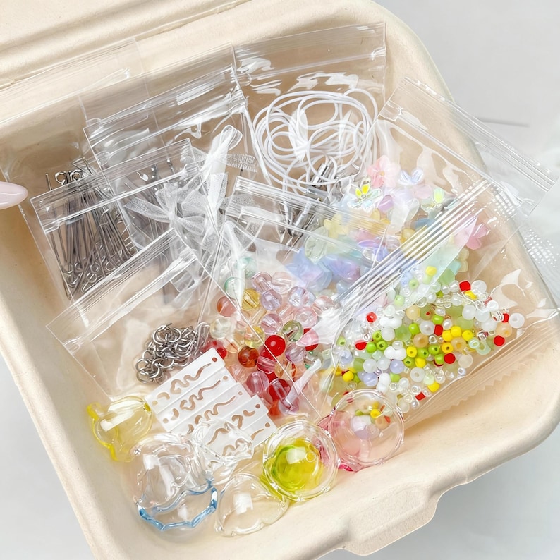 May include: A craft kit with assorted beads, charms, and findings in clear plastic bags. The kit includes colourful beads, small metal pins, and clear glass-like components. The items are arranged in a light brown container.