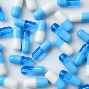 Puede incluir: Vista cenital de numerosas cápsulas azules y blancas dispersas sobre una superficie blanca. Las cápsulas son cilíndricas, con un cuerpo azul claro o blanco y una tapa de color contrastante. La disposición es aleatoria, creando un patrón de color y forma.