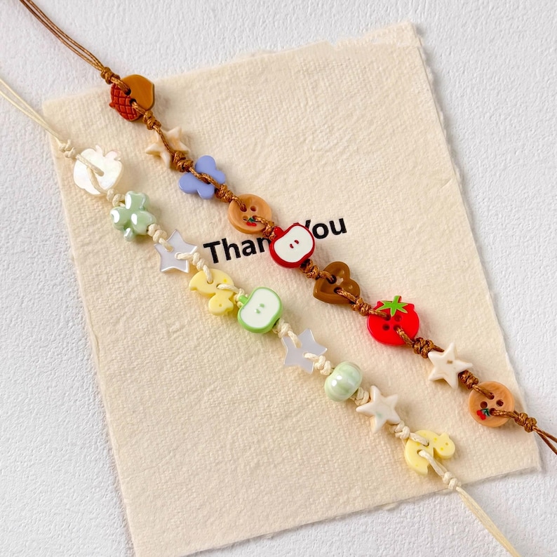 May include: Two handmade beaded bracelets with fruit and star charms. One bracelet features a red strawberry, a green apple, and yellow lemon charms. The other bracelet has a red apple charm. The background has the words "Thank you".