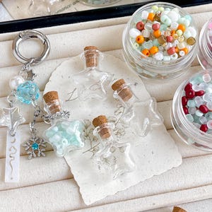 May include: A collection of craft supplies featuring small glass star-shaped bottles with cork stoppers, a keychain with charms, and small jars filled with colorful beads. The bottles are clear, and the beads are in various colors.