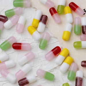 Puede incluir: Un conjunto de cuentas coloridas en forma de cápsula en varios colores, incluyendo blanco, rosa, verde, amarillo y rojo. Las cuentas están dispersas sobre una superficie texturizada blanquecina, creando una exhibición vibrante. Ideales para la creación de joyas.