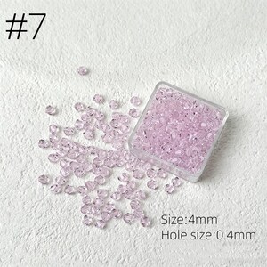 May include: A collection of light purple crystal beads, 4mm in size, with a 0.4mm hole, are scattered on a white surface. Some beads are contained in a clear square container. The number "7" is in the upper left corner.