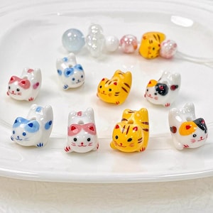 Może przedstawiać: Kolekcja ceramicznych koralików w kształcie kota w różnych kolorach, w tym białym, żółtym i kaliko. Koraliki mają niebieskie, różowe i pomarańczowe akcenty. Niektóre koraliki są nawleczone na przezroczyste i różowe koraliki. Koraliki leżą na białym talerzu.