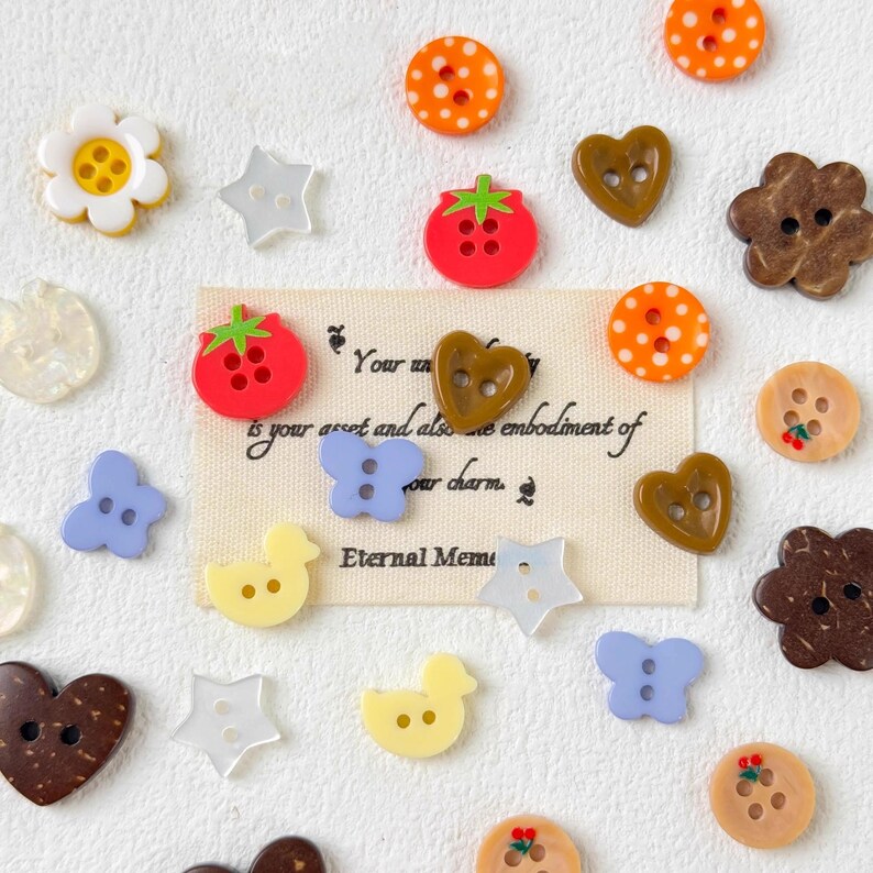 May include: A collection of decorative buttons in various shapes and colors, including flowers, stars, hearts, and ducks. Some buttons are red with green accents, others are orange with white polka dots. A small card with text is also present.