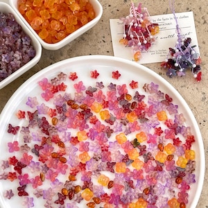 Könnte beinhalten: Eine Sammlung bunter Blüten- und Schmetterlingsperlen in Orange-, Rosa-, Rot- und Lilatönen. Einige Perlen sind auf einem weißen Teller verstreut, andere in kleinen weißen Behältern. Zwei Perlenornamente sind ebenfalls zu sehen, zusammen mit einer handschriftlichen Notiz.