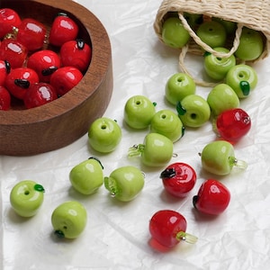 Pode incluir: Contas de vidro em forma de maçã em vermelho e verde. Algumas estão numa tigela de madeira, outras espalhadas numa superfície branca e outras a sair de um pequeno cesto de vime. As contas têm um acabamento brilhante.