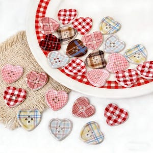 May include: A collection of heart-shaped buttons in various plaid patterns, including red, pink, brown, and blue. The buttons are arranged on a white plate with a red and white checkered rim and a beige fabric.