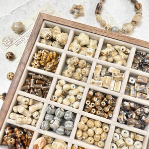 May include: A wooden tray containing an assortment of beige and brown ceramic beads in diverse shapes and sizes. The beads include star, round, and cylindrical forms. A matching beaded bracelet is also present.