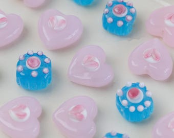 May include: Close-up of a collection of decorative glass beads. The beads are in the shape of hearts and small squares. The heart-shaped beads are light pink with a white floral design. The square beads are blue with white and pink accents.