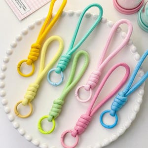 Può includere: Una collezione di portachiavi colorati nei toni del giallo, verde, rosa e blu. Ogni portachiavi presenta un anello di corda annodata e un moschettone in metallo abbinato. I portachiavi sono disposti su un piatto bianco con bordo smerlato.