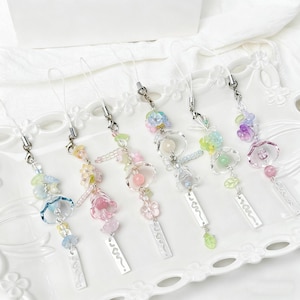May include: Six decorative phone charms with clear straps and silver-tone clasps. Each charm features pastel-coloured beads and flower-shaped charms. Each charm has a small, rectangular tag. The charms are displayed on a white, decorative plate.