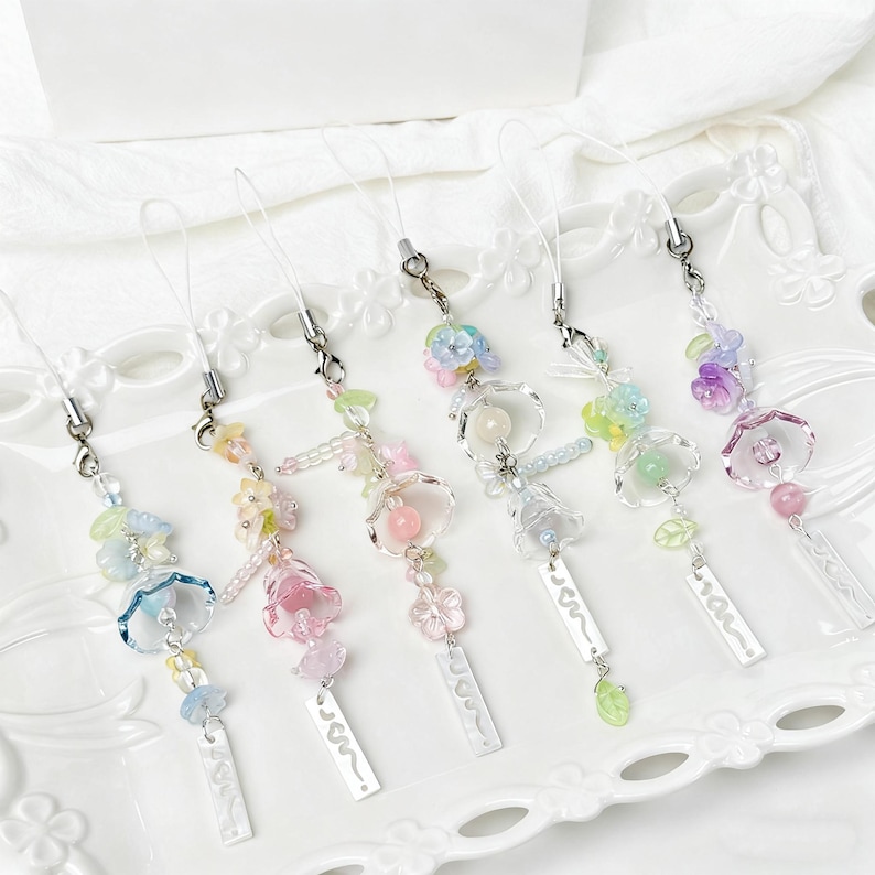 May include: Six decorative phone charms with clear straps and silver-tone clasps. Each charm features pastel-coloured beads and flower-shaped charms. Each charm has a small, rectangular tag. The charms are displayed on a white, decorative plate.