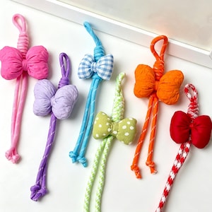 Può includere: Una collezione di accessori per capelli a forma di fiocco colorati. Ogni fiocco è realizzato in tessuto e attaccato a un cordino abbinato. I fiocchi sono disponibili in vari colori e fantasie, tra cui rosa, viola, quadretti blu, verde a pois bianchi, arancione e rosso.