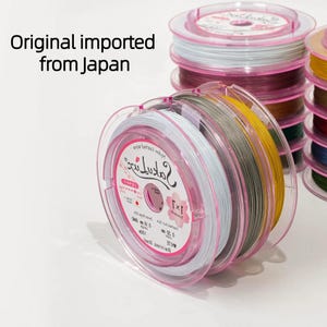 Puede incluir: Carretes de hilo de pescar en varios colores, incluyendo blanco, gris y amarillo, se muestran en estuches de plástico rosa transparente. El texto "Original imported from Japan" es visible. Los carretes están apilados, mostrando los colores.