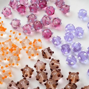 May include: A collection of colorful glass beads in various shades of pink, orange, brown, and purple. The beads have a textured, spiky appearance, and some feature white polka dots. These beads are ideal for jewelry making and crafting.