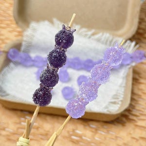 Puede incluir: Dos brochetas de caramelos morados con forma de bayas. Una brocheta tiene caramelos morados oscuros, la otra caramelos morados claros. Los caramelos están en brochetas de madera, con algunos caramelos y una caja en el fondo.