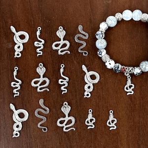 May include: Silver snake charms in different poses, with a beaded bracelet featuring a snake charm. The charms show scale detail and coiled or slithering designs. The bracelet has white, grey, and black beads. The background is a wooden surface.