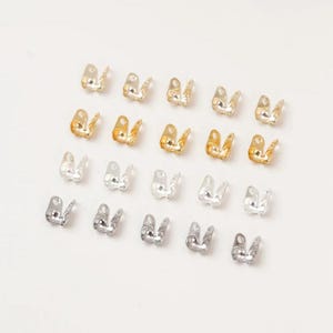 May include: An assortment of small, metal crimp end findings in gold and silver tones. These jewelry-making components are arranged on a white surface. The findings are designed to secure the ends of cords or ribbons.