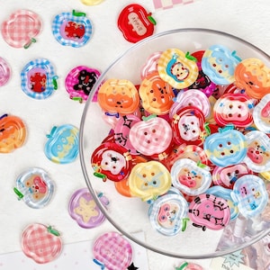 Puede incluir: Una colección de coloridos botones con forma de manzana en un cuenco de cristal transparente y sobre una superficie blanca. Los botones presentan varios patrones y diseños, incluyendo cuadros, rayas y personajes de dibujos animados. Algunos tienen la palabra "APPLE".