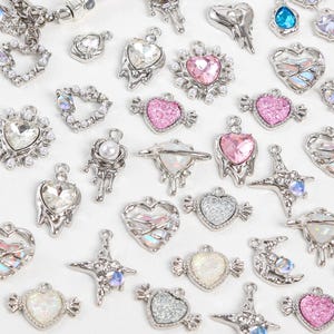 May include: A collection of silver-toned charms in various shapes, including hearts, stars, and moons. Some charms feature clear or pink gemstones, pearls, and iridescent accents. The charms are designed for jewelry making or crafting.