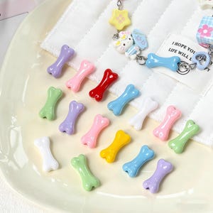 May include: A selection of colourful, bone-shaped charms in shades of purple, pink, red, blue, white, yellow, and green. The charms are displayed on a pale yellow plate, with white fabric in the background. Some charms are part of a keychain.