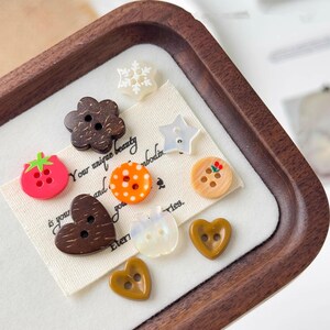 May include: A collection of decorative buttons in various shapes and colors, including a strawberry, flower, star, and heart. The buttons are arranged on a white fabric with text and displayed in a wooden tray.