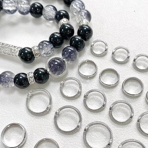 Op de afbeelding: Een assortiment sieraden, waaronder armbanden met kralen en zilveren ringen. De armbanden hebben zwarte en heldere kralen, met zilveren accenten. De ringen zijn zilverkleurig, sommige met een textuurontwerp. De sieraden worden op een wit oppervlak gepresenteerd.