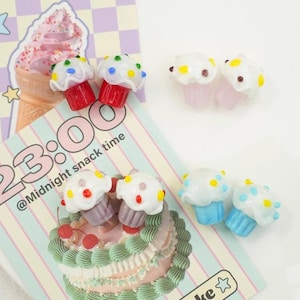 以下が含まれることがあります： 白いアイシングとカラフルなスプリンクルが付いた、ミニチュアカップケーキ型のチャームのコレクション。 カップケーキのベースは赤、ピンク、紫、青です。 画像には、「23:00 @Midnight snack time」というテキストが書かれた装飾カードも含まれています。