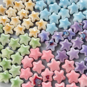 May include: A selection of small, star-shaped ceramic beads in a spectrum of colours. The beads vary from light yellow to pink, green, blue, and purple. Each star has five points and a glossy finish, ideal for jewellery making or crafts.
