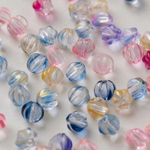 May include: A collection of translucent, faceted beads in various colors, including pink, blue, yellow, and purple. The beads have a swirled, striped design and are scattered across a white surface. These beads are suitable for jewelry making and crafts.
