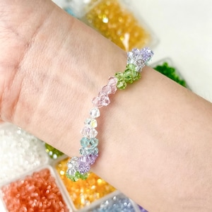 May include: A colorful beaded bracelet with a gradient of colors, including green, pink, purple, and blue. The bracelet is made of small, faceted beads, and is worn on a wrist. The background shows containers of colorful beads.