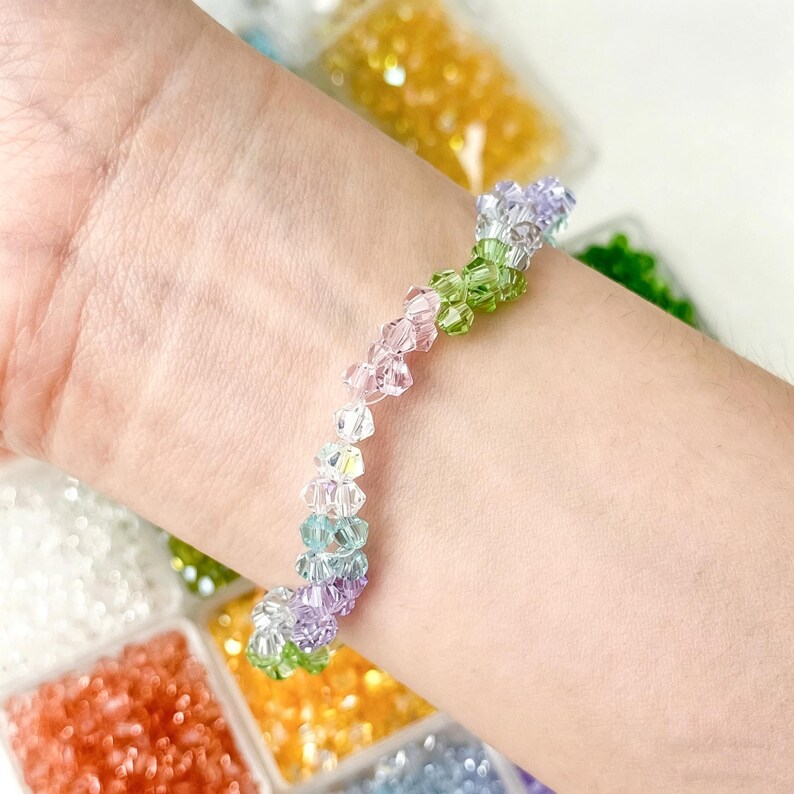 May include: A colorful beaded bracelet with a gradient of colors, including green, pink, purple, and blue. The bracelet is made of small, faceted beads, and is worn on a wrist. The background shows containers of colorful beads.