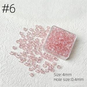 May include: Pink crystal beads, size 4mm, are scattered on a white surface, with some contained in a clear plastic square container. The hole size is 0.4mm. The number 6 is in the upper left corner.