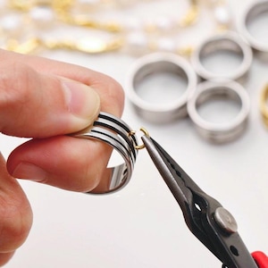以下が含まれることがあります： 黒いストライプが入ったシルバーリングを手に持ち、小さなペンチを使ってゴールドカラーのリングを取り付けています。他のシルバーリングとゴールドのネックレスが背景にあります。