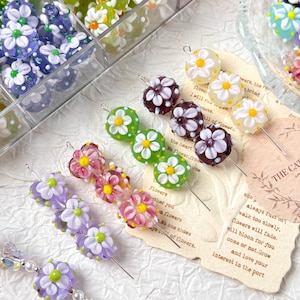May include: A selection of colourful glass flower beads in shades of purple, pink, green, and white. The beads are on metal pins, ideal for jewellery making. Some beads are stored in a clear plastic container.
