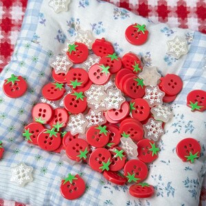 Puede incluir: Una colección de botones rojos con forma de tomate con tallos verdes y botones transparentes con forma de copo de nieve. Los botones están esparcidos sobre una tela estampada floral azul y blanca, con un fondo a cuadros rojos y blancos.
