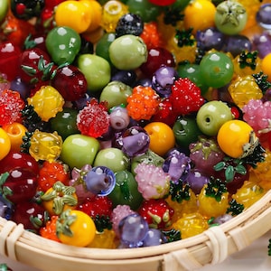 Puede incluir: Una colección de cuentas de frutas de vidrio de colores, que incluyen manzanas, uvas, fresas y piñas. Las cuentas son en tonos de verde, rojo, amarillo, naranja y morado. Las cuentas están en una cesta tejida.