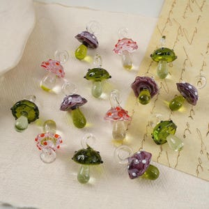 May include: A selection of miniature glass mushroom charms. The caps are in shades of pink, purple, and green, some with white or red spots. Each charm has a clear stem and a loop for attachment.