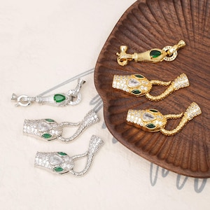 Puede incluir: Una variedad de cierres de joyería en tonos dorados y plateados, con diseños de cabeza de serpiente. Los cierres están adornados con piedras preciosas claras y verdes, y se exhiben en una bandeja de madera.