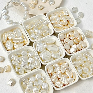 May include: An assortment of off-white, irregular-shaped pearls and beads. The pearls are displayed in small, square white dishes, with some loose pearls scattered around. A pearl necklace is visible in the upper left corner.