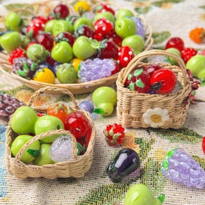 May include: Miniature glass fruit sculptures, including apples, strawberries, and grapes, are displayed in woven baskets and a round dish. The fruits are in various colours, such as red, green, and purple. The baskets have handles and are decorated with a flower.