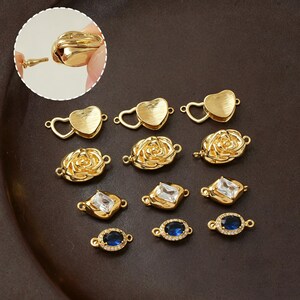 May include: A collection of gold-toned jewelry connectors featuring various designs including heart shapes with brushed textures, three-dimensional rose motifs, rectangular clear crystals, and oval blue stones surrounded by small white accents.