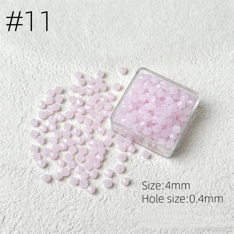 May include: A collection of light pink faceted beads, scattered on a white surface, with some contained in a clear plastic box. The text "#11" is in the upper left corner. The text indicates the beads are 4mm in size with a 0.4mm hole.