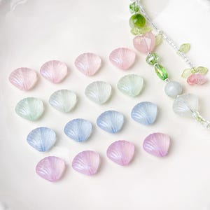 May include: A collection of iridescent seashell-shaped beads in pastel colors, including pink, blue, green, and lavender. A beaded strand with green, pink, and clear beads is also visible. The beads are arranged on a white surface, ideal for jewelry making or craft projects.