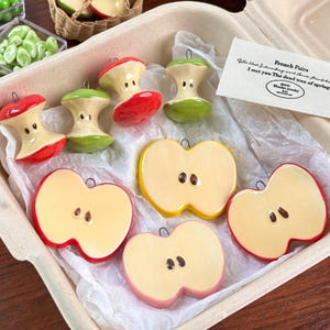 Puede incluir: Una colección de adornos de cerámica con forma de manzana en un recipiente de cartón. Los adornos tienen forma de mitades y corazones de manzana, con detalles rojos, amarillos y verdes. Una pequeña etiqueta dice "French Pairs".
